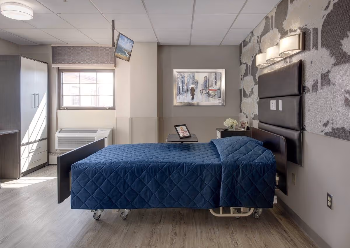 A neatly made hospital-style bed with a dark blue quilted blanket in a well-lit room. The room features a large window with natural light, a wall-mounted TV, a wardrobe, a nightstand with a flower vase and framed photo, and a painting on the wall. The wall behind the bed has a patterned wallpaper and three modern light fixtures.