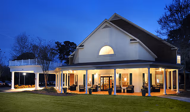 Exterior view of a senior living facility building at dusk with warm lights glowing from inside. The building has a large covered porch with seating areas and white columns, surrounded by a well-maintained lawn and trees in the background.