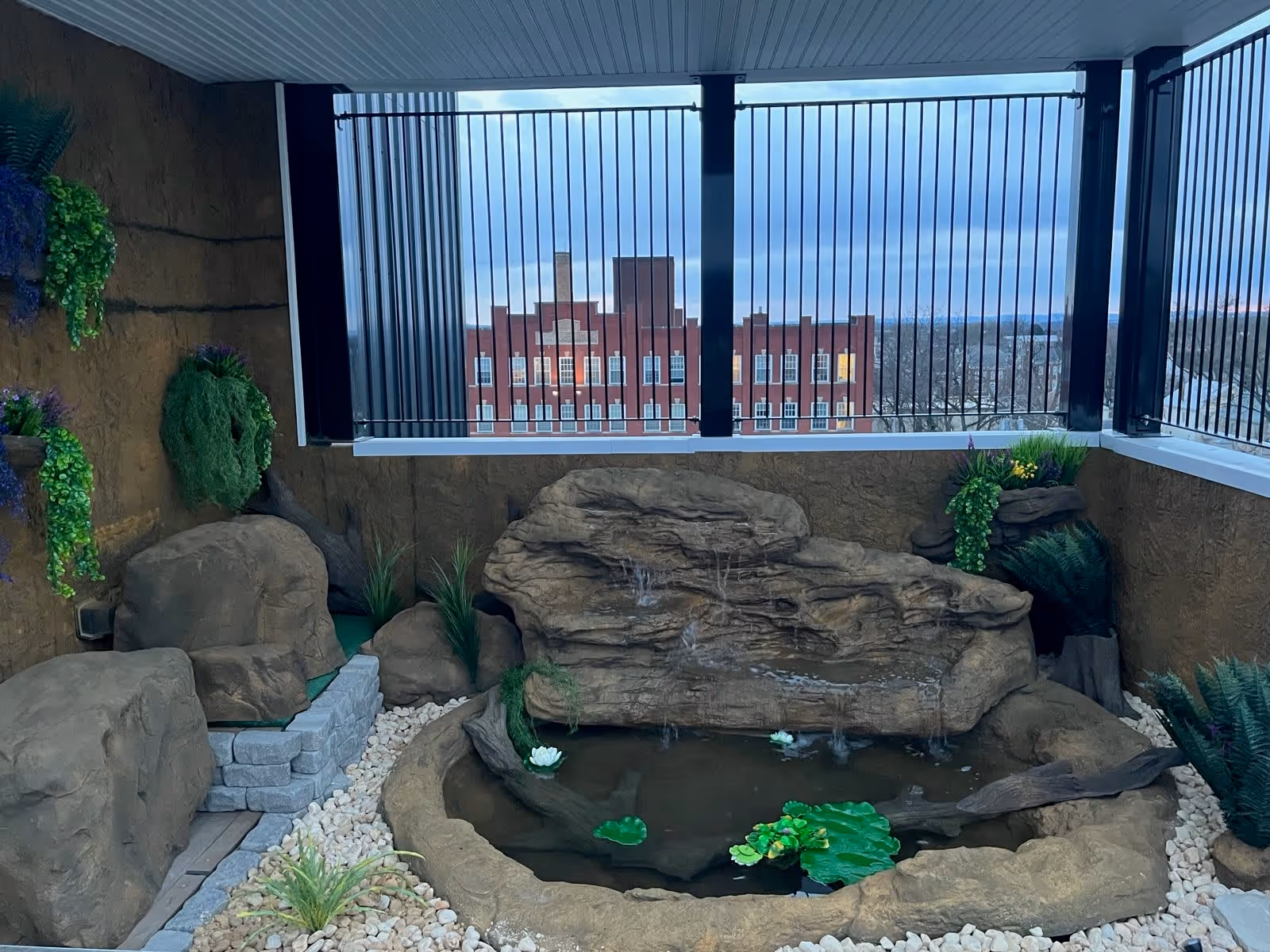 An outdoor balcony area with a small artificial rock waterfall and pond surrounded by decorative plants and stones. The balcony has black vertical railings and offers a view of a red brick building and a cloudy sky in the background.