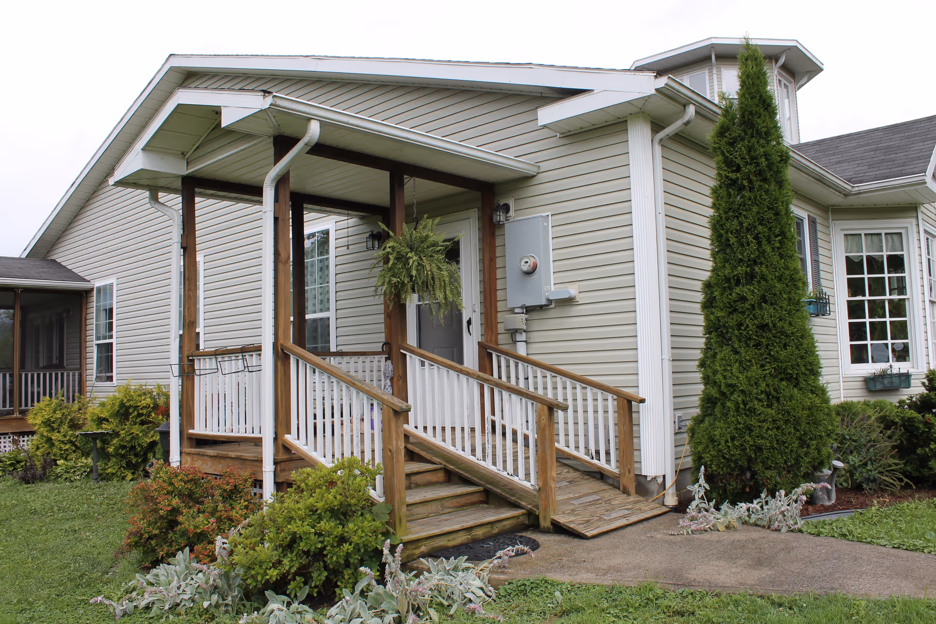 Exterior view of a single-story building with beige siding and a small covered porch with wooden railings and steps leading up to a white door. There is a hanging plant on the porch and various shrubs and greenery surrounding the building. A tall evergreen tree is planted near the corner of the building.