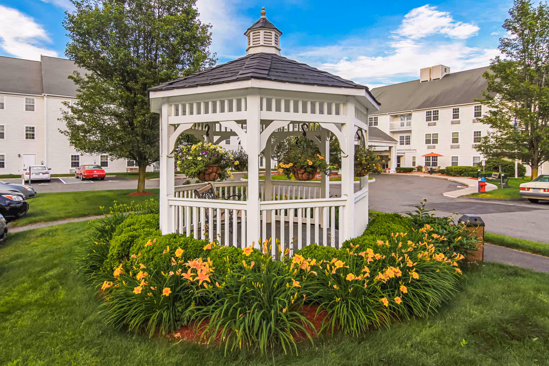 A white wooden gazebo surrounded by blooming orange and yellow flowers and green bushes, situated on a well-maintained lawn with trees and a paved driveway leading to a large white multi-story building in the background under a partly cloudy blue sky.