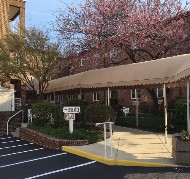 Outdoor entrance area of a brick building with a covered walkway, surrounded by greenery and blooming trees. There are signs indicating the entrance and a parking area with marked spaces.