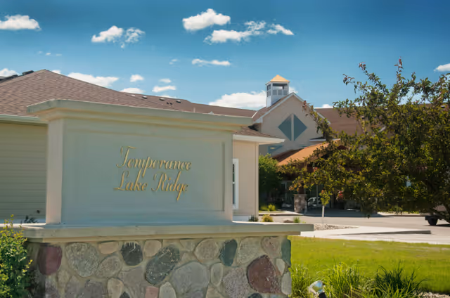Stone and concrete entrance sign reading "Temperance Lake Ridge" with the senior living building and landscaping behind it under a blue sky.