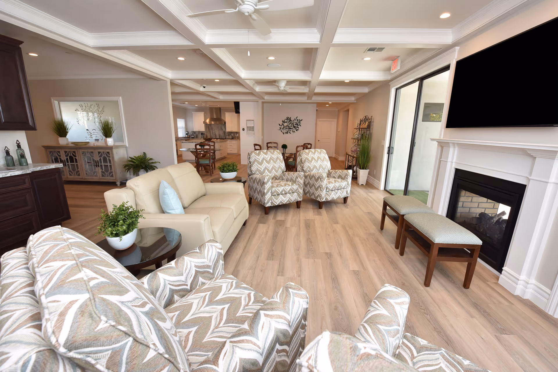 A bright and spacious living room area in a senior living facility with patterned armchairs, a beige leather sofa, a fireplace with a mounted TV above it, and a view into a dining area and kitchen in the background. The room features light wood flooring, recessed lighting, ceiling fans, and decorative plants.
