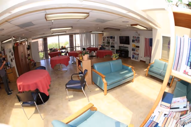 A bright common area in a senior living facility with several tables covered in red tablecloths, blue cushioned chairs and sofas, bookshelves filled with books, a television mounted on the wall, and large windows letting in natural light.