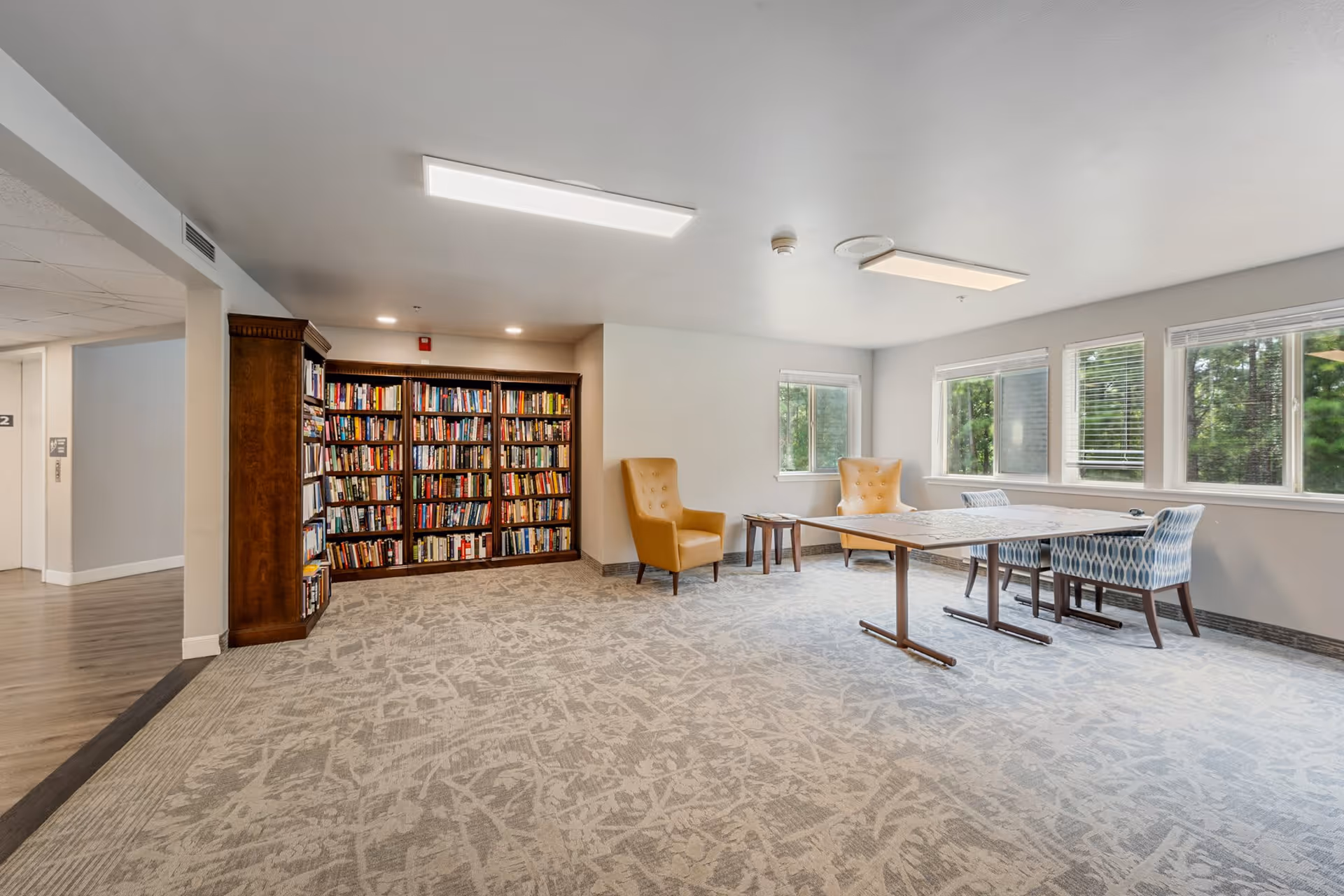 A bright and spacious room with large windows showing greenery outside. The room features a wooden bookshelf filled with books, two mustard yellow armchairs, a wooden table with a puzzle on it, and two patterned blue chairs. The floor is carpeted with a light patterned design, and the walls are painted light gray.