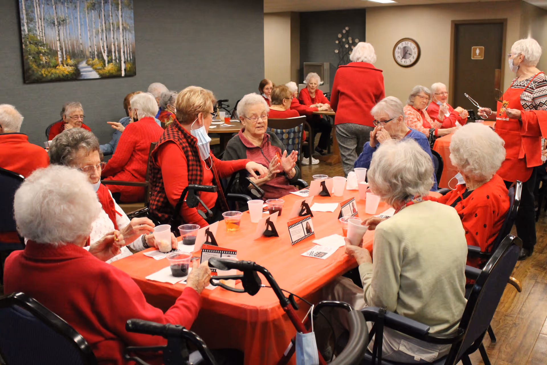 A group of elderly residents seated around tables with red tablecloths socializing and having drinks in a common dining area.