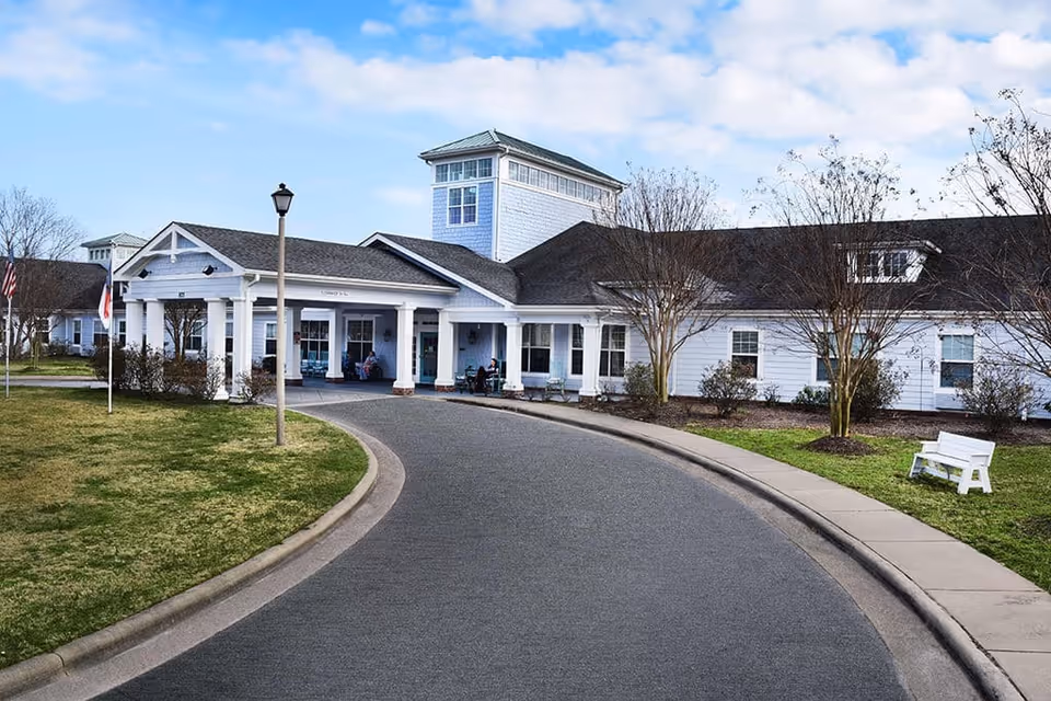 Front entrance and circular driveway of a white senior living facility with a covered portico, columns, and surrounding lawn.