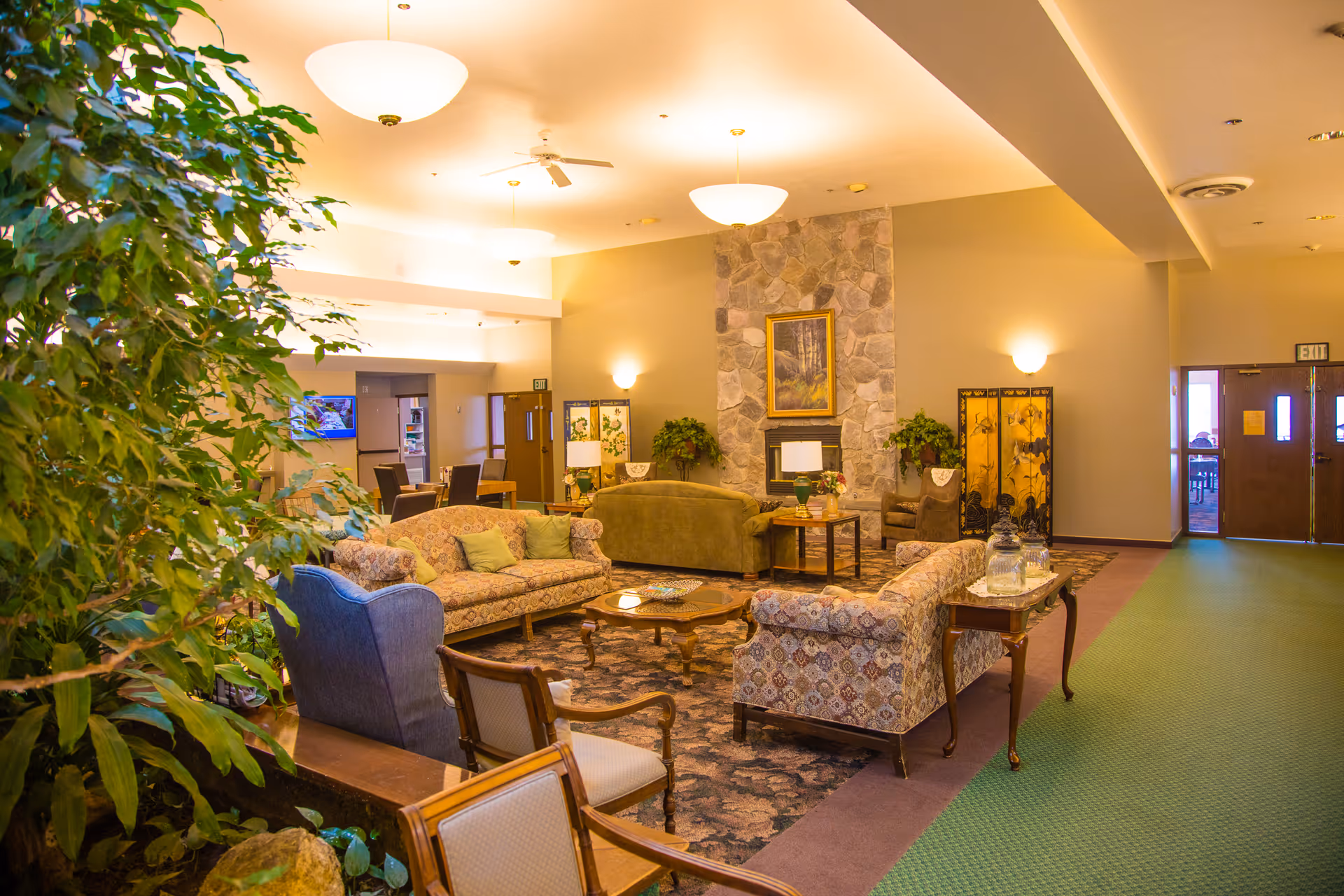 A cozy assisted living facility common area with patterned sofas, armchairs, a stone fireplace with a framed painting above it, side tables with lamps, and indoor plants. The room has warm lighting and a green carpeted floor with a floral patterned rug in the seating area. There are exit doors and a TV mounted on the wall in the background.