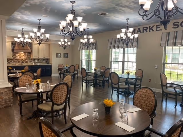 Communal dining room with round wooden tables, upholstered chairs, chandeliers, and large windows.