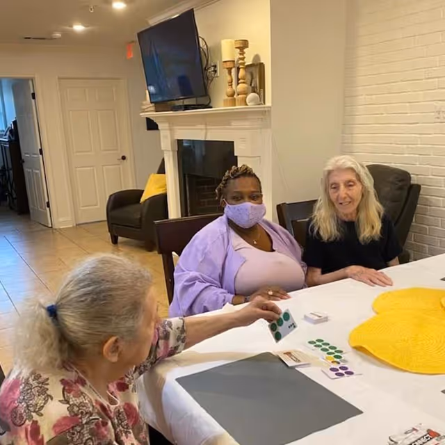 Three women sitting around a table in a living room setting. One woman is wearing a purple mask and purple clothing, another woman with long white hair is sitting next to her, and a third woman with gray hair tied back is reaching across the table holding a card with green dots. The table has a white tablecloth, some cards, and a large yellow hat. Behind them is a fireplace with a TV mounted above it and decorative items on the mantel.