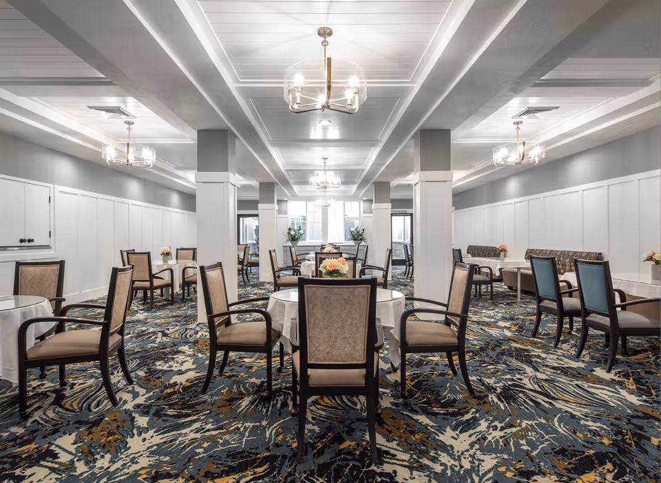 Bright formal dining room with round tables, upholstered chairs, chandeliers, and a patterned carpet.