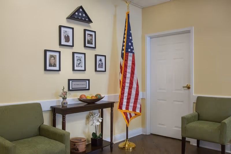 A small seating area with two green armchairs facing each other, a wooden table between them holding a decorative bowl with colorful balls and a small vase with flowers. Above the table is a display titled 'The Wall of Honor' featuring five framed black and white portraits and a folded American flag in a triangular case. An American flag on a stand is positioned next to a closed white door in the corner of the room with light yellow walls and dark wood flooring.