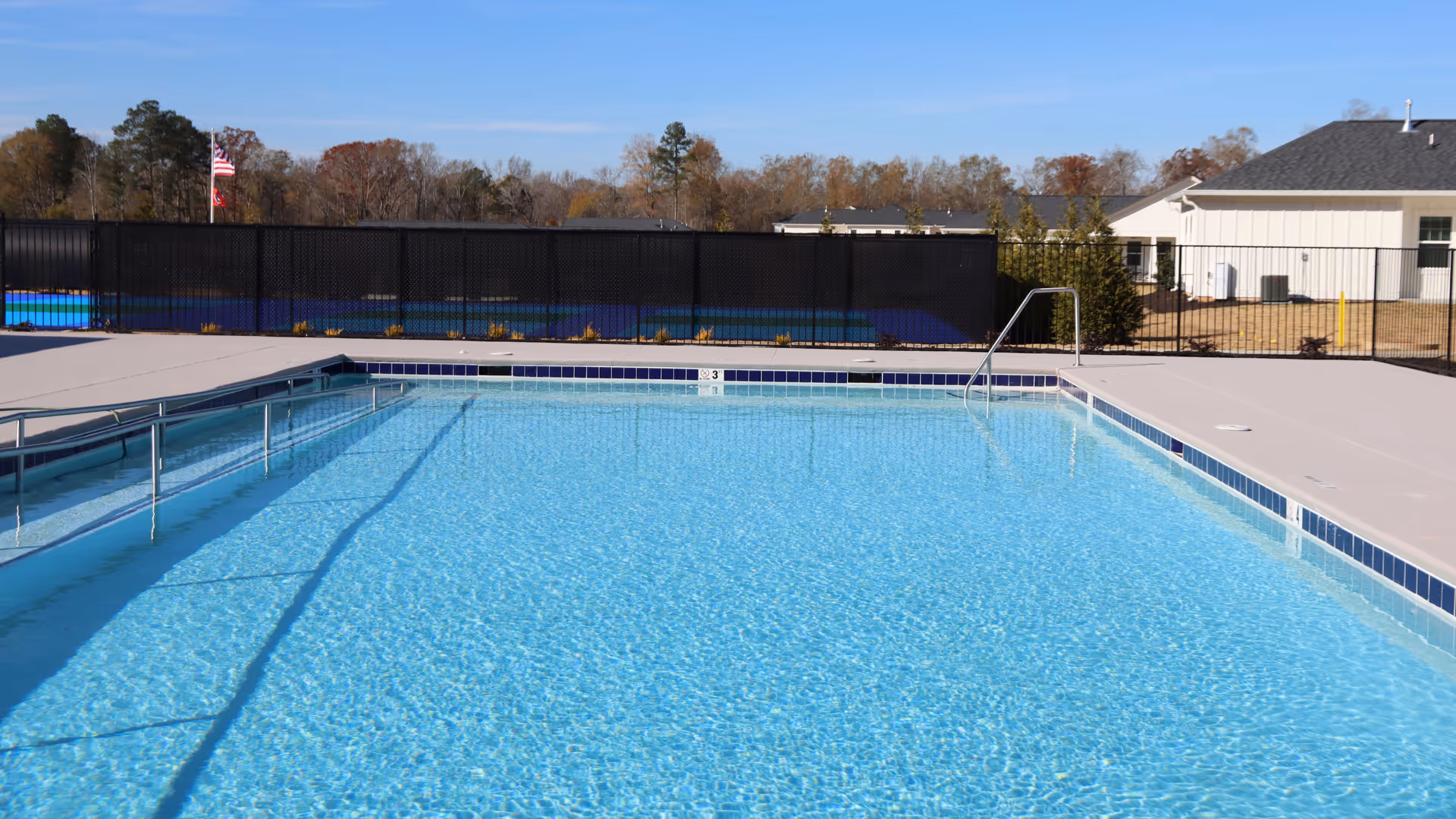 Outdoor swimming pool with clear blue water, surrounded by a concrete deck and a black safety fence. In the background, there are trees, a building, and an American flag on a flagpole under a clear blue sky.