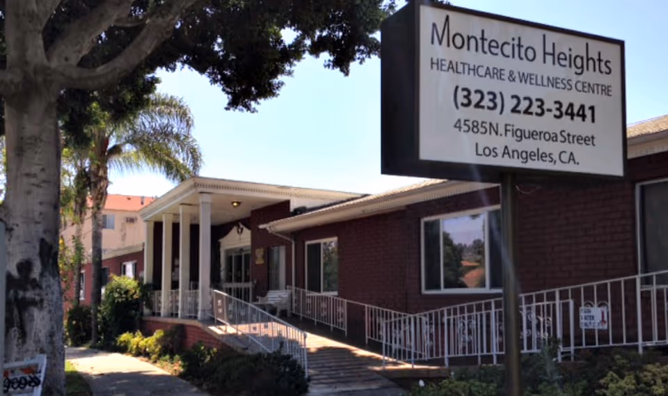 Exterior view of Montecito Heights Healthcare & Wellness Centre building with a large sign displaying the facility name, phone number, and address. The building has a brick facade, a ramp with white railings, and is surrounded by trees and plants.
