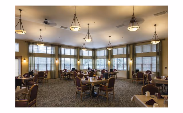 A spacious dining room with multiple tables and chairs arranged neatly. Each table is set with napkins, cups, and condiments. Large windows with green curtains allow natural light to fill the room. Several ceiling lights hang from above, illuminating the area.