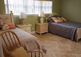 A bedroom with two single beds, each with a bedside table and lamp. The bed on the left has a beige patterned bedspread and a decorative pillow, while the bed on the right has a dark gray bedspread with multiple pillows. There is a wooden chair with a cushion in the foreground, and a large window with vertical blinds letting in natural light. The walls are painted light green and the floor is tiled.