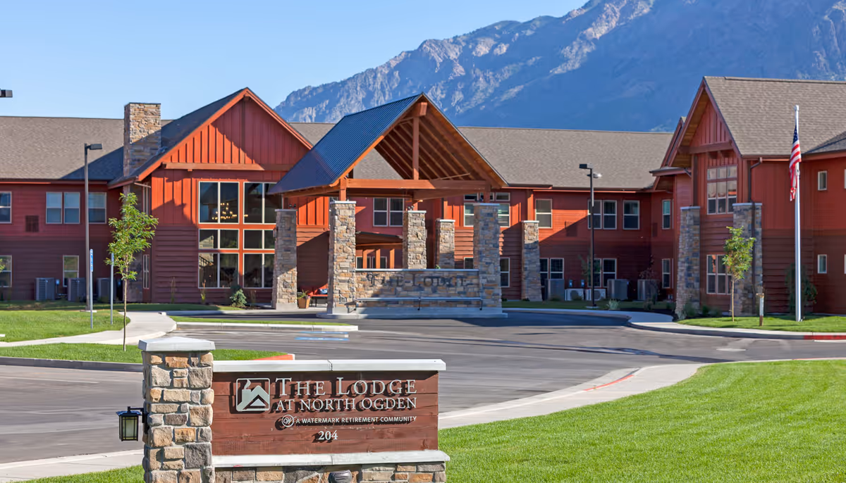 Exterior view of The Lodge at North Ogden, a large red building with stone pillars and a covered entrance, set against a backdrop of mountains under a clear blue sky. There is a sign in the foreground with the facility name and address, surrounded by green grass and a paved driveway.