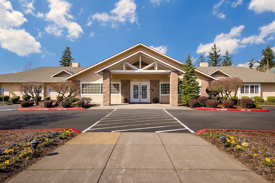 Single-story senior living building with a covered central entrance, landscaped shrubs, and a parking/drop-off area under a blue sky.