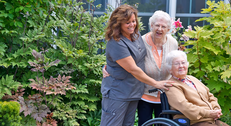 A smiling caregiver in gray scrubs stands outdoors with two elderly women, one seated in a wheelchair and the other standing beside her, surrounded by lush green plants and flowers.