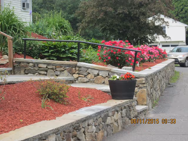 Outdoor garden area with stone retaining walls, a black metal handrail, red mulch, green plants, and vibrant pink flowers. A black planter with red and white flowers is placed on the stone wall. A driveway and a white vehicle are visible in the background.