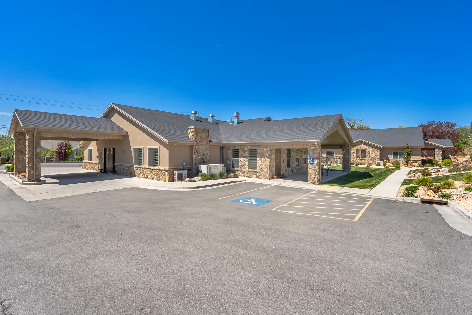Exterior view of a single-story senior living facility building with stone and beige stucco walls, a covered entrance, and a parking lot with a designated handicapped parking space. The sky is clear and blue, and there is landscaping with grass, bushes, and rocks around the building.