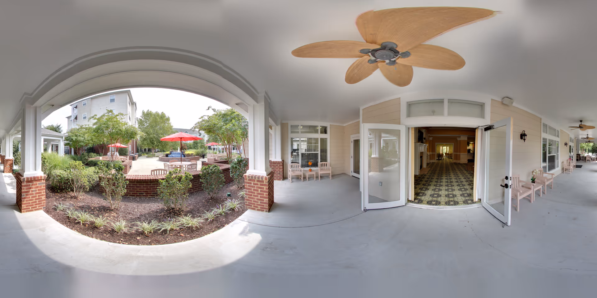 Covered outdoor walkway with ceiling fans and seating, leading to an open entrance of a building. Outside the walkway, there is a garden area with shrubs and trees, and a patio with tables and red umbrellas.