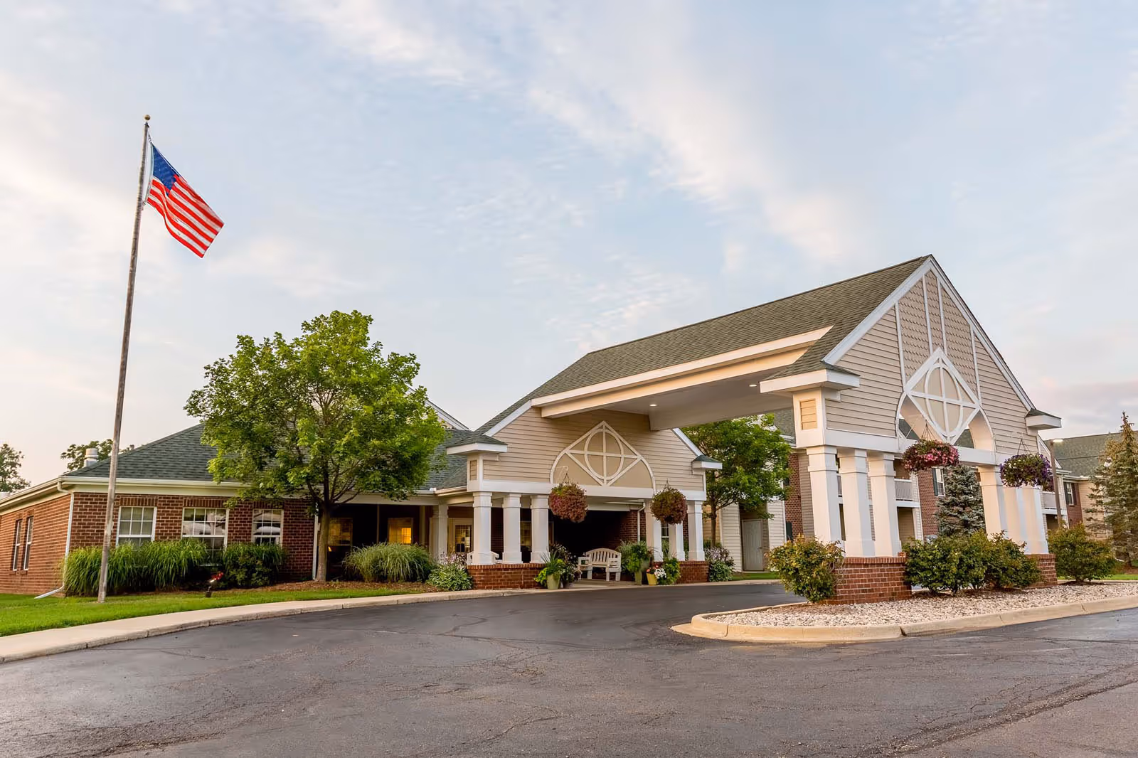 Exterior view of Independence Village of Grand Ledge showing the entrance with a covered driveway, brick and siding building, hanging flower baskets, green trees, and an American flag on a flagpole.