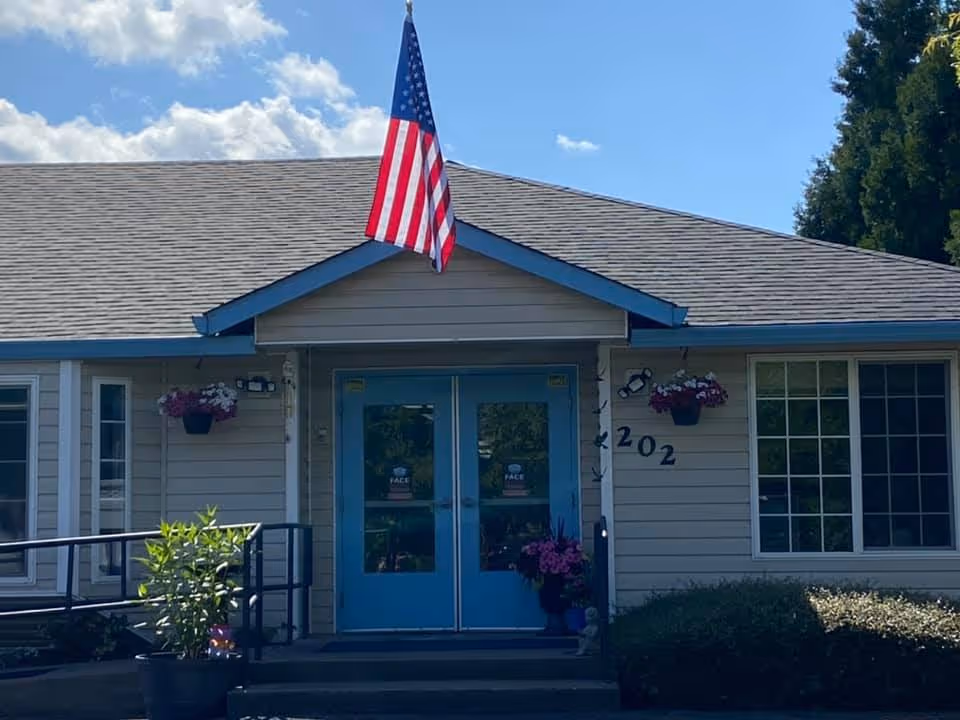 Front exterior view of a single-story building with beige siding and a gray shingled roof. The entrance features double blue doors with glass panels, flanked by hanging flower pots with pink and white flowers. An American flag is mounted above the entrance. The building number 202 is displayed on the right side of the doors. There are windows on either side of the entrance and some greenery in front of the building.