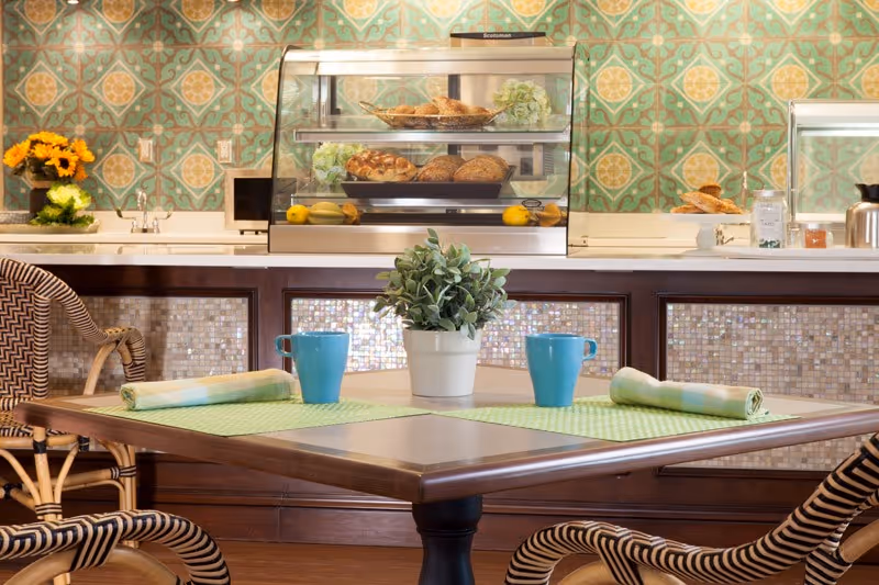 A cozy dining area with a wooden table set for two, featuring green placemats, rolled napkins, and blue mugs. In the background, there is a counter with a glass display case containing various breads and pastries, a vase with sunflowers, and a decorative green and yellow patterned wall.