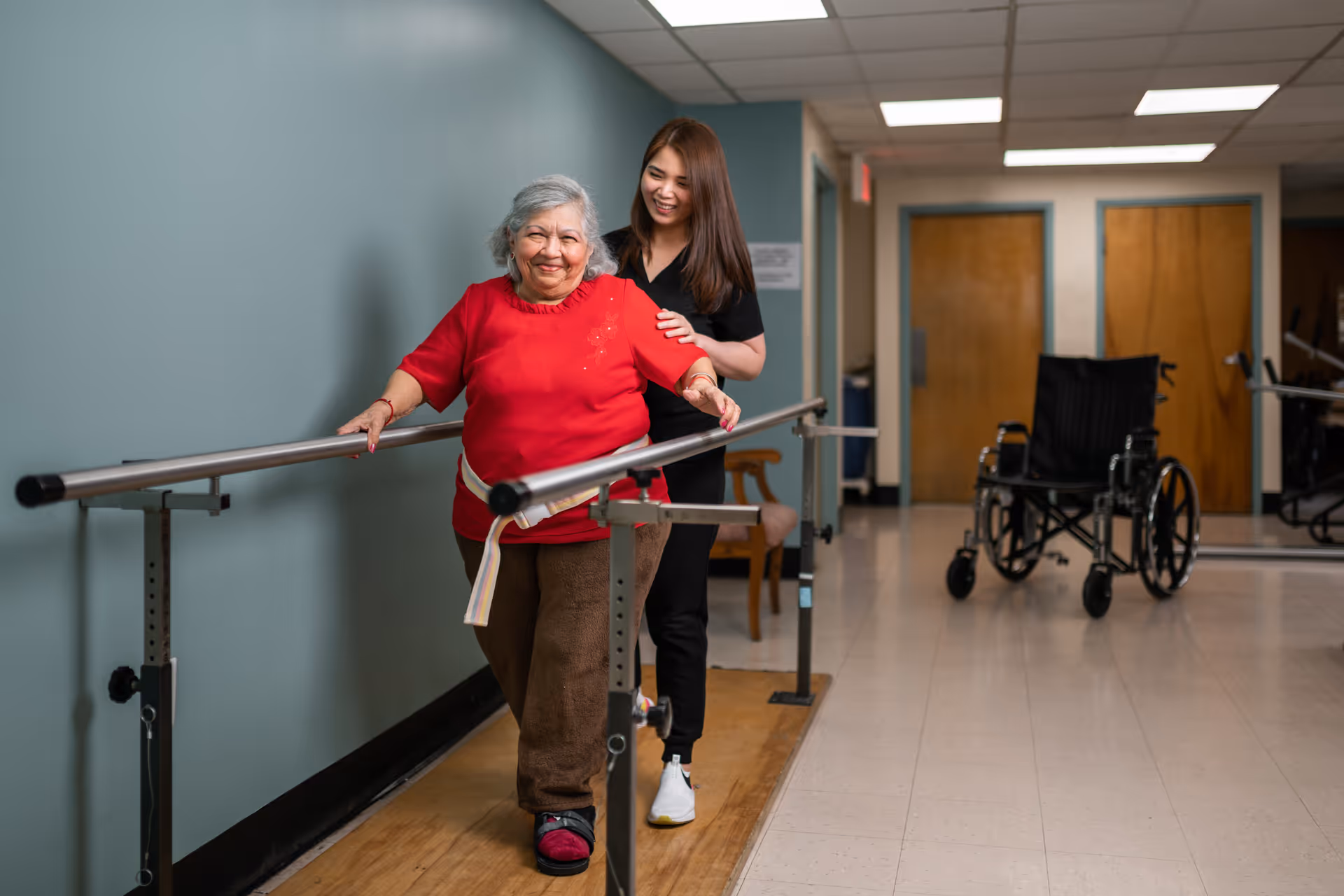 An elderly woman in a red sweater is walking with support from parallel bars in a rehabilitation center hallway, assisted by a smiling caregiver dressed in black. A wheelchair is visible in the background near wooden doors.