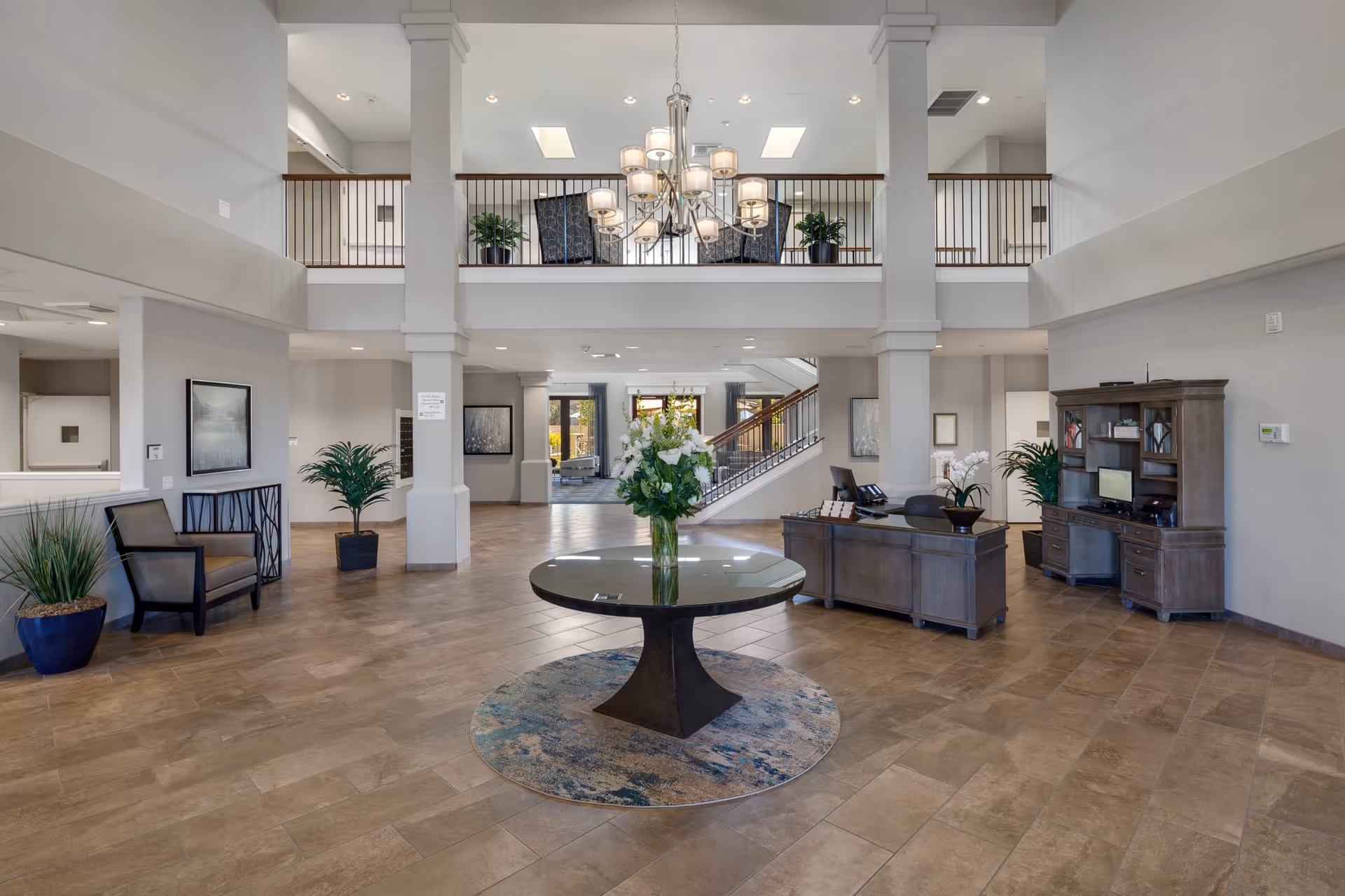 Spacious and well-lit senior living facility lobby with a round table featuring a flower arrangement in the center, a chandelier hanging from the ceiling, seating areas with chairs and plants, a reception desk, and a staircase leading to an upper floor with a balcony.