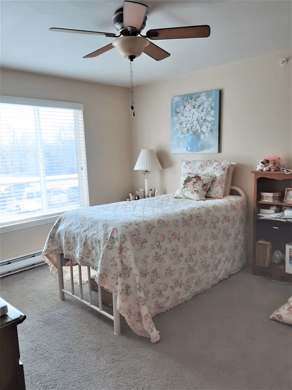 Sunlit bedroom with a floral-covered single bed, bedside lamp, ceiling fan, painting on the wall, and a window with blinds.