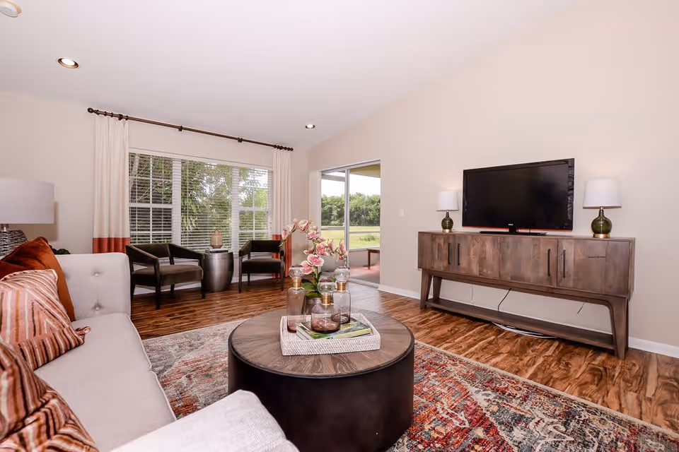 A bright living room with a beige sofa adorned with striped and solid rust-colored pillows, a round wooden coffee table with decorative items, two dark armchairs near a large window with white and rust-colored curtains, a wooden TV console with a flat-screen TV and two green table lamps, and a colorful patterned rug on a wooden floor. A sliding glass door opens to an outdoor area with greenery visible.