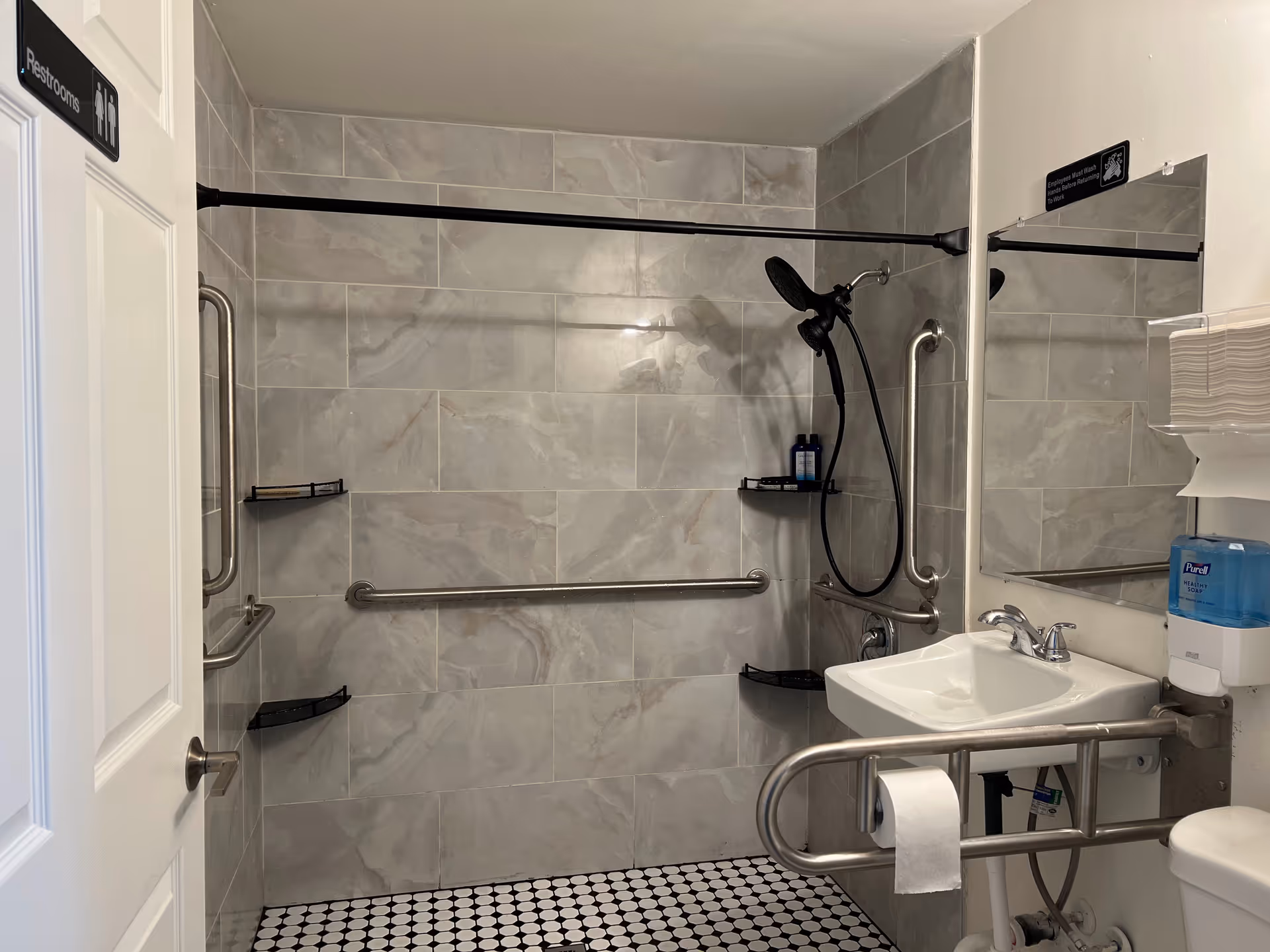 Accessible bathroom with gray tiled walls and black and white tiled floor. The bathroom features a wall-mounted sink with grab bars, a toilet with a grab bar and toilet paper holder, a shower area with a handheld showerhead, multiple grab bars, and corner shelves holding toiletries. A mirror and a soap dispenser are mounted on the wall above the sink. The door has a sign indicating restrooms.