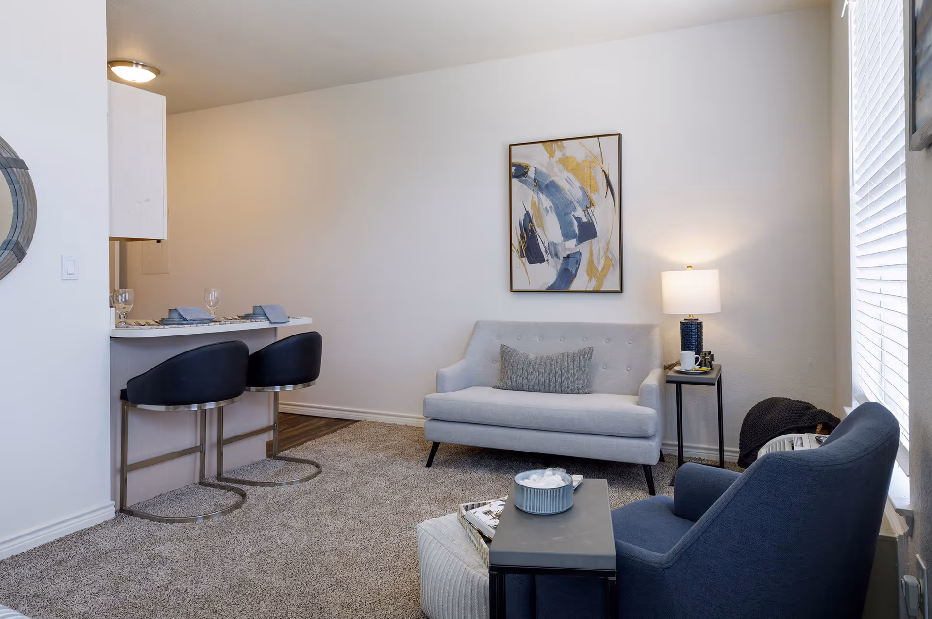 A cozy living room area with a light gray loveseat, a dark blue armchair, a small side table with a lamp, and a coffee table with decorative items. There is a modern abstract painting on the wall and a window with blinds letting in natural light. To the left, there is a breakfast bar with two black bar stools and place settings.