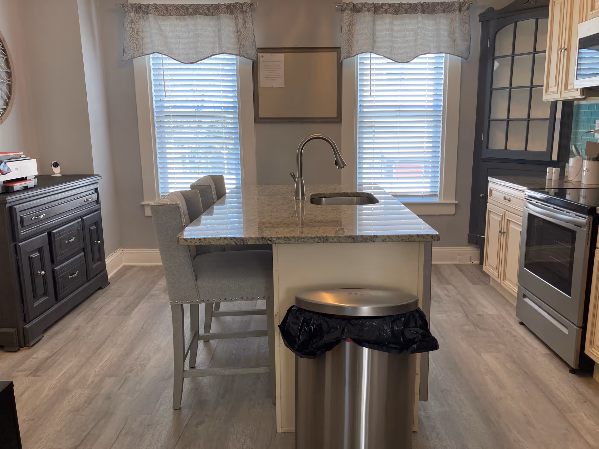 A modern kitchen with a central island featuring a granite countertop and a built-in sink with a curved faucet. Three upholstered bar stools are positioned along one side of the island. The kitchen has light-colored cabinets, a stainless steel oven, and a stainless steel trash can with a black liner in front of the island. Two windows with blinds and decorative valances allow natural light into the room. A dark wooden cabinet is visible on the left side of the image.