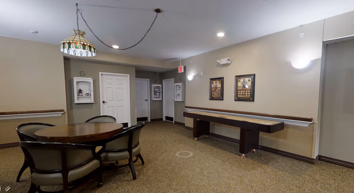 A common area in a senior living facility featuring a round wooden table with four cushioned chairs, a shuffleboard table against the wall, beige walls with two framed playing card artworks, and multiple white doors. The room is carpeted and has ceiling lights and a stained glass hanging lamp.