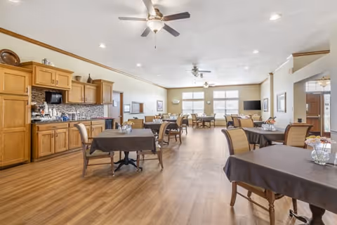 Bright communal dining area with multiple tables and chairs, a kitchenette with wood cabinets, ceiling fans, and large windows.