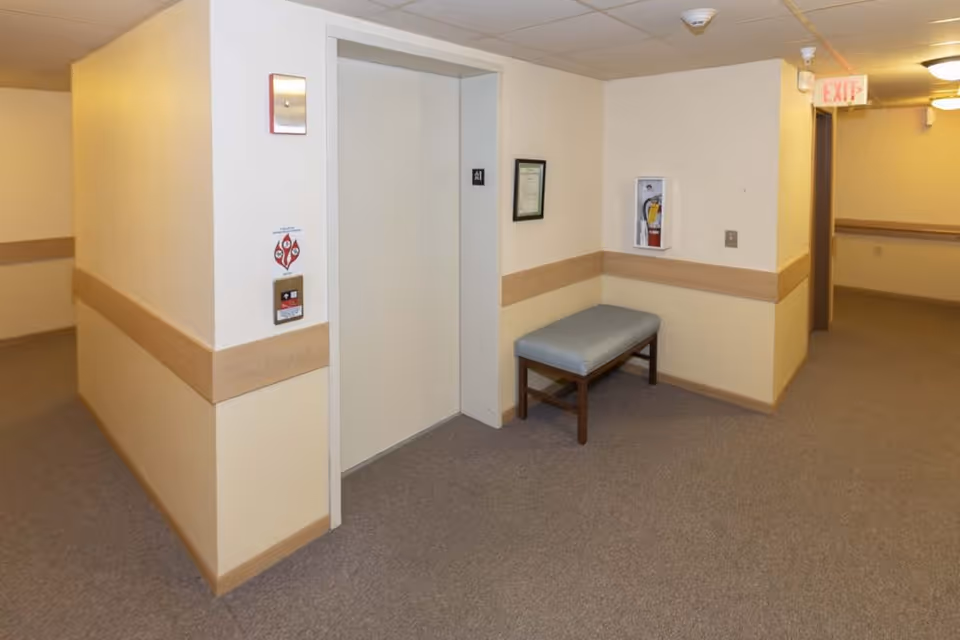 Interior hallway of McFarland Villa Assisted Living featuring a closed elevator door, a small bench with a cushion, a fire extinguisher mounted on the wall, beige walls with wooden trim, carpeted floor, and an exit sign above a doorway in the background.
