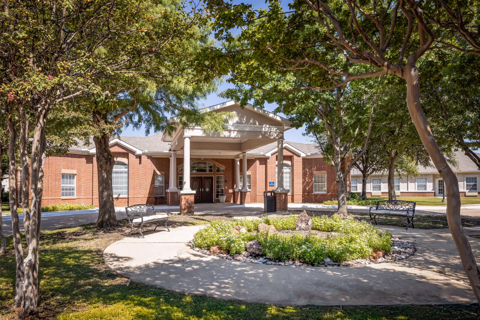 Front entrance of a red-brick senior living building with columns, a circular landscaped driveway, benches, and shade trees.