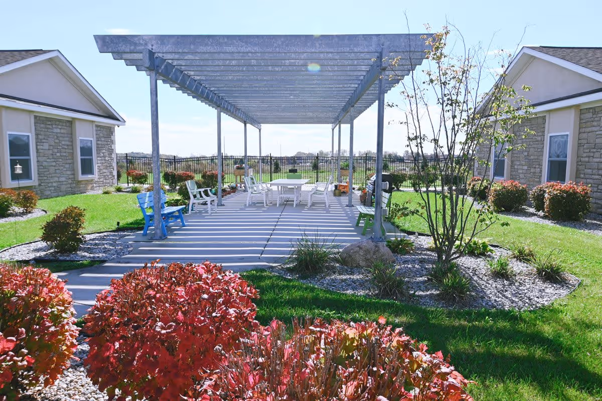 Outdoor patio area with a pergola providing partial shade over a concrete seating area with white tables and chairs. The patio is surrounded by green grass, red and green bushes, small trees, and two single-story buildings with stone and beige siding on either side.