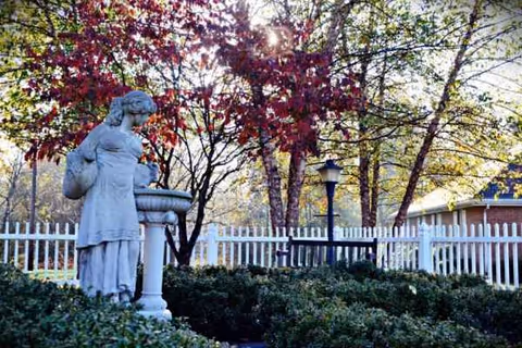 Stone statue beside a fountain in a garden with a white picket fence.