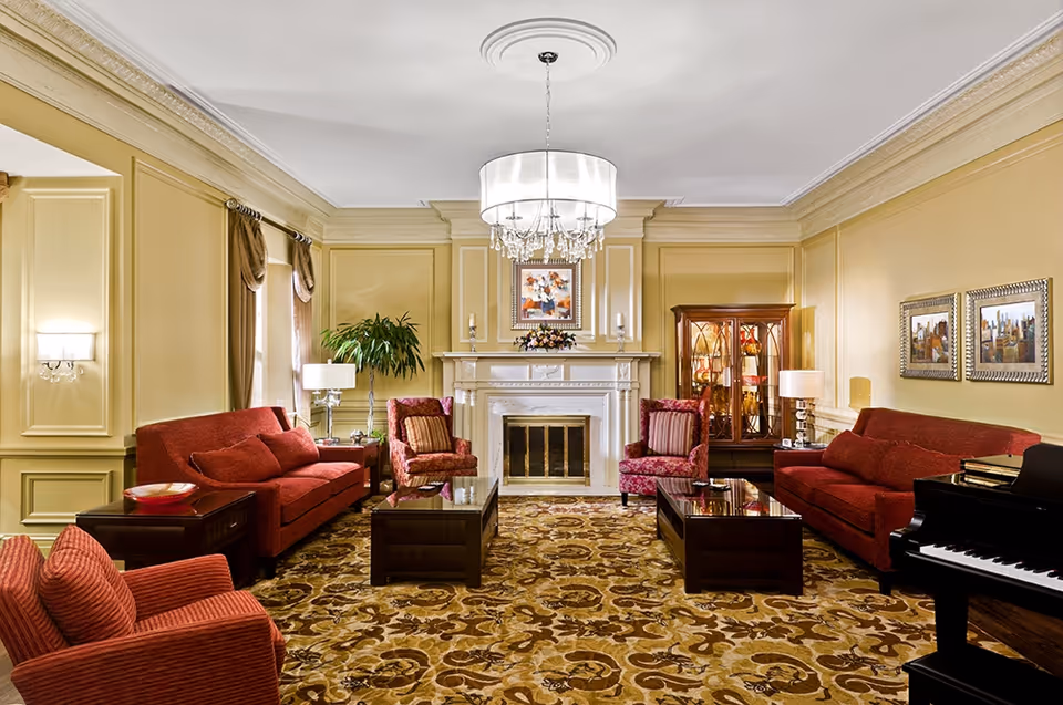 A spacious and elegant living room with beige walls and a patterned carpet. The room features two red sofas, two red armchairs, two dark wood coffee tables, a white fireplace with decorative items on the mantel, a chandelier hanging from the ceiling, a glass display cabinet, framed artwork on the walls, and a black grand piano on the right side.