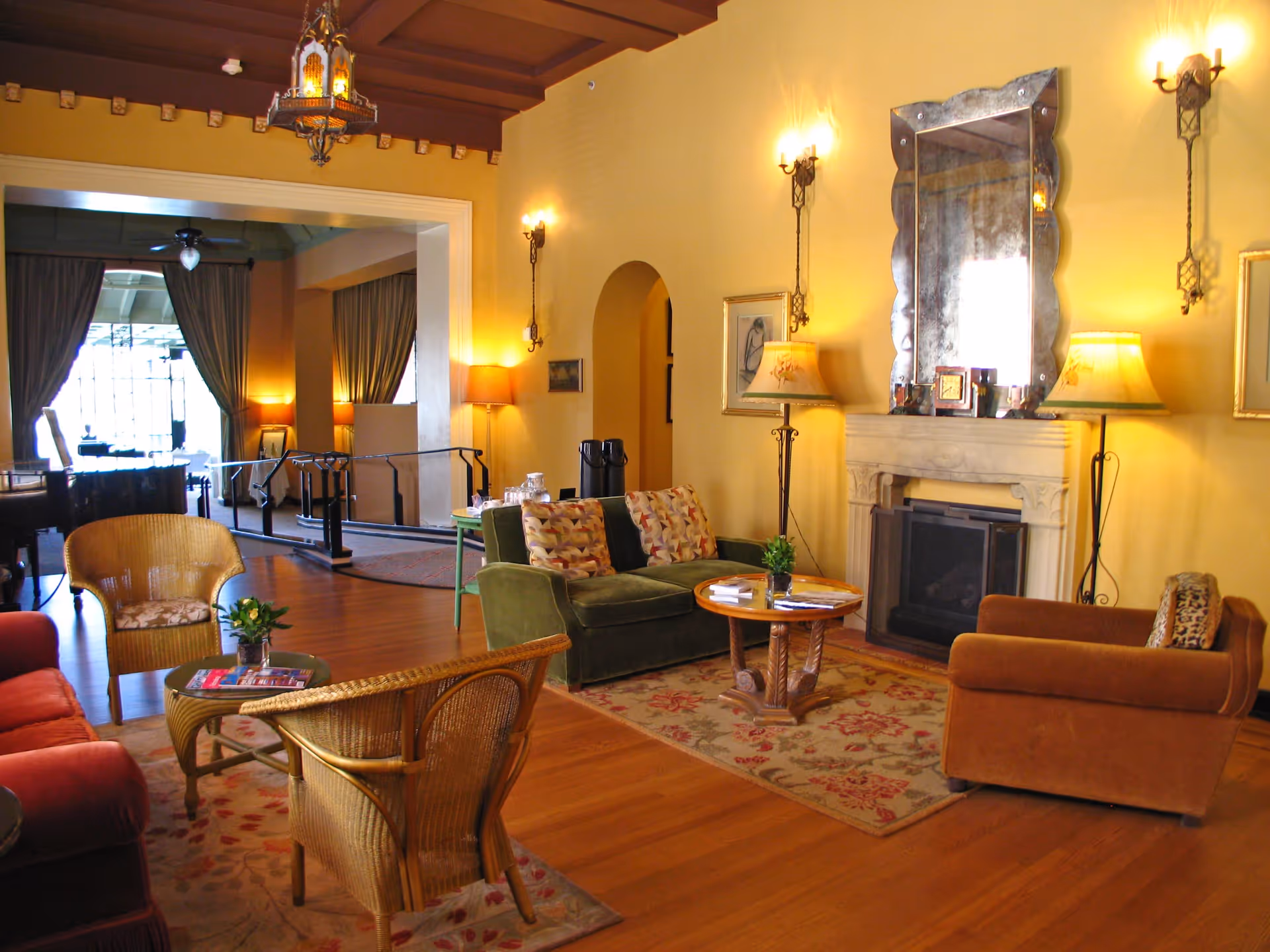 A cozy living room with wooden floors, a red sofa, two wicker chairs, a green loveseat, and a brown armchair arranged around two small wooden tables. The room features a fireplace with a large decorative mirror above it, two floor lamps, wall sconces, and a hanging lantern-style light fixture. In the background, there is an open doorway leading to another room with large windows and curtains.