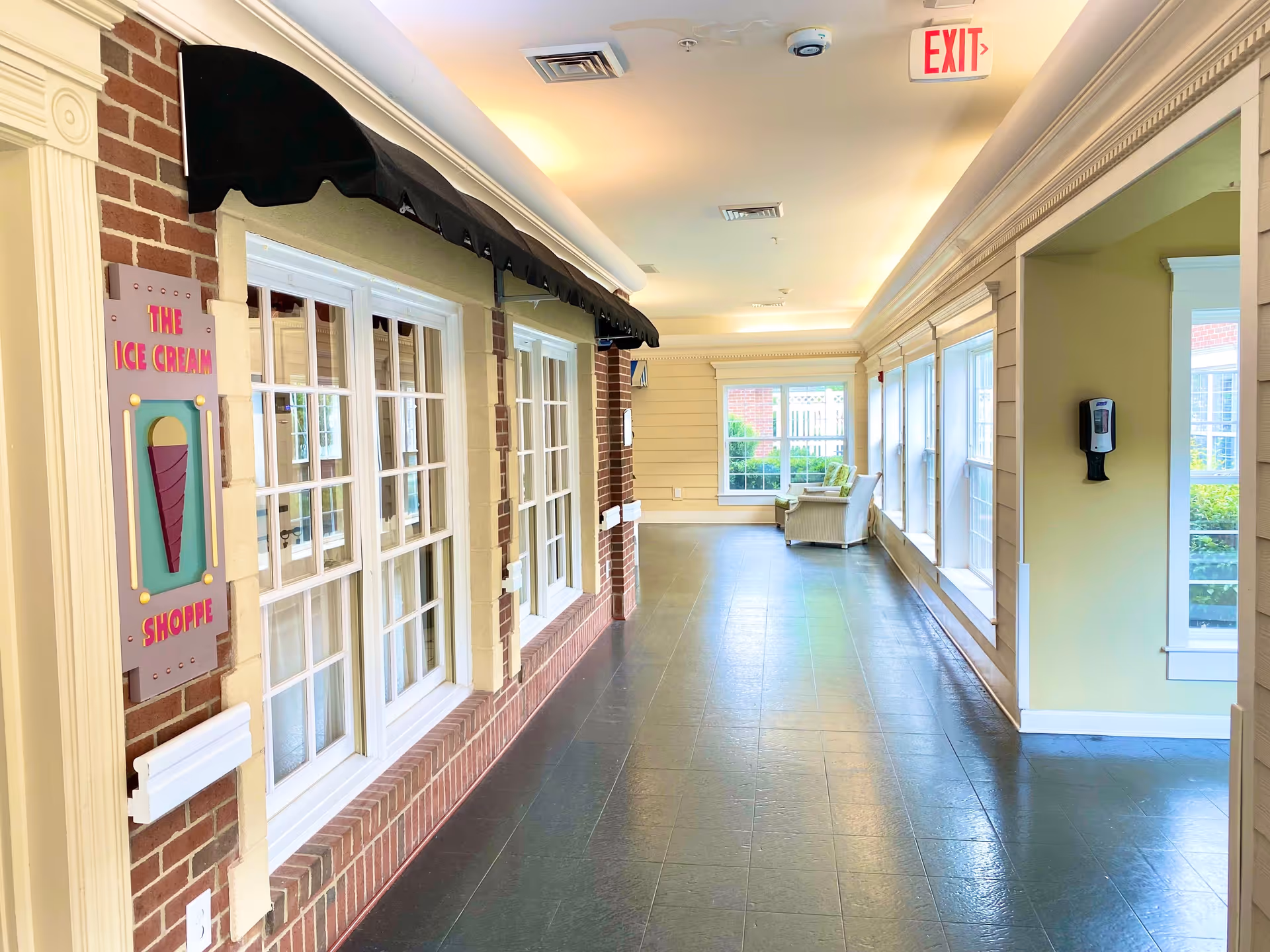 Indoor hallway with large windows on the right side and a brick wall on the left featuring a sign that reads 'The Ice Cream Shoppe' with an ice cream cone graphic. The hallway has a tiled floor, a black awning above the windows on the left, and a seating area with chairs at the far end near a window. An exit sign is visible on the ceiling.