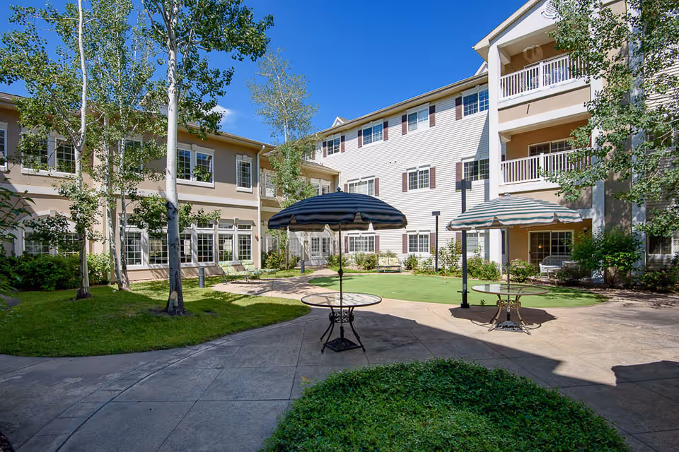 Outdoor courtyard area of The Inn At Garden Plaza with patio tables and umbrellas, surrounded by a three-story building with balconies and windows, green grass, trees, and clear blue sky.