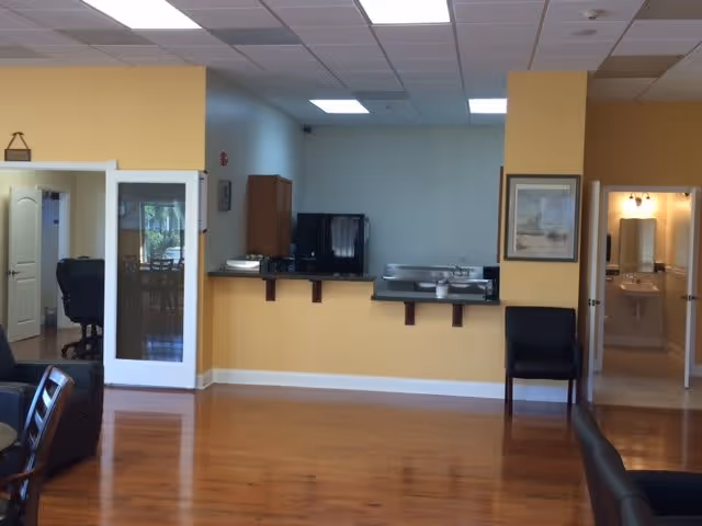 Interior view of a senior living facility common area with wooden flooring, yellow and white walls, a counter with a coffee maker and sink, a black chair, and an open doorway leading to a bathroom with a sink and mirror.