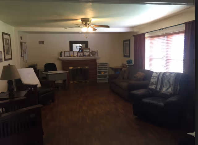 A living room with wooden flooring, a ceiling fan with lights, a brick fireplace with a mantle displaying framed pictures and decorations, a desk with a chair, a brown sofa with cushions, a black armchair with a patterned throw, and windows with blinds and curtains letting in natural light.