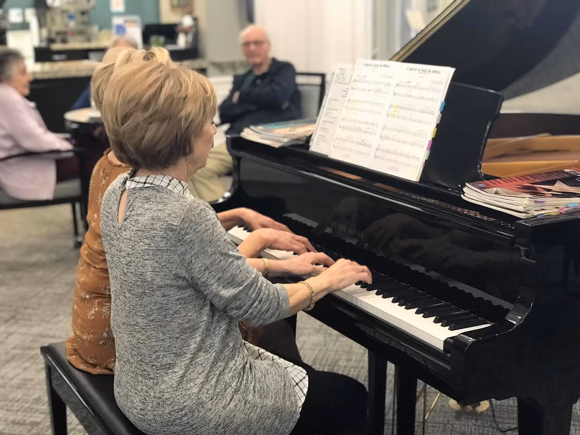 Two elderly women playing a black grand piano together in a common area, with sheet music on the piano and other elderly people sitting and watching in the background.
